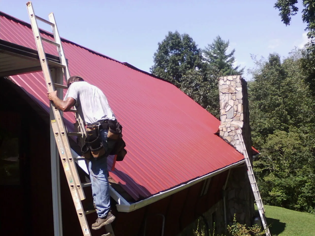 Roofer on a ladder beside a red metal roof and stone chimney — Soffit & Fascia Repair by Roofing Brothers in Lake City